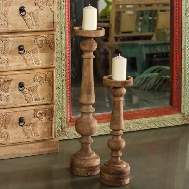 Two tall wooden candle holders with a rustic finish, each holding a candle, placed in front of a decorative mirror.