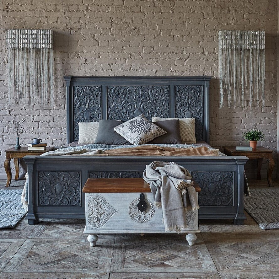 Decorative bedroom with an ornate gray bed and wooden chest against a brick wall.